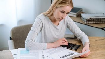 Une femme concentrée travaille, en utilisant une calculatrice et consultant plusieurs documents étalés devant elle.