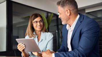 Deux professionnels discutent dans un bureau. La femme tient une tablette et sourit à l'homme en costume bleu.