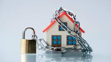 Une petite maison miniature en plastique est entourée par une chaîne en métal, fermée par un cadenas doré.