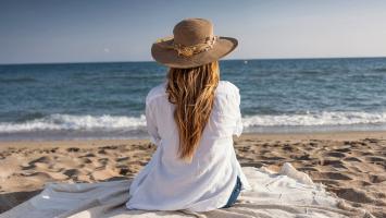 Une femme assise sur une couverture à la plage, regardant la mer, portant un chapeau de paille et une chemise blanche.