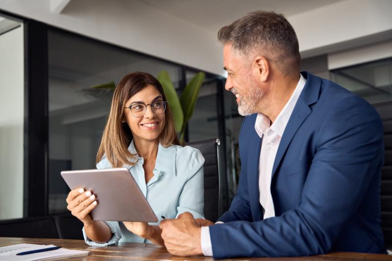 Deux professionnels discutent dans un bureau. La femme tient une tablette et sourit à l'homme en costume bleu.