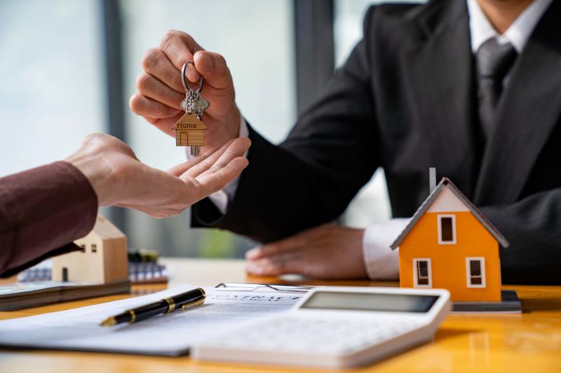 Un agent immobilier remet les clés d'une maison à un nouveau propriétaire, symbolisé par une maquette de maison et un contrat sur une table