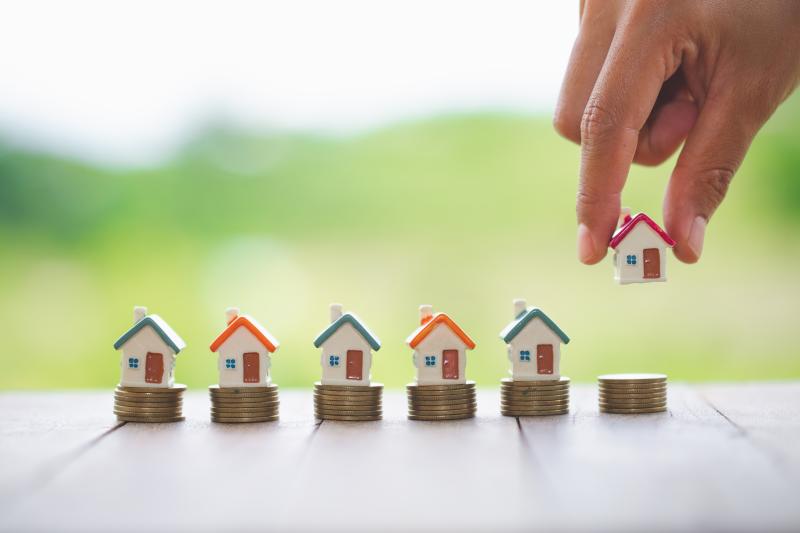 Une main place une maison miniature sur des pièces, avec quatre maisons similaires en arrière-plan.