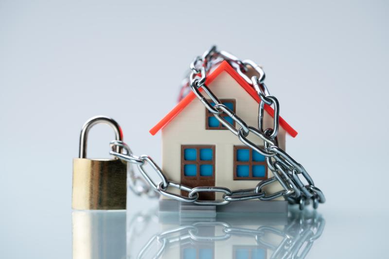 Une petite maison miniature en plastique est entourée par une chaîne en métal, fermée par un cadenas doré.