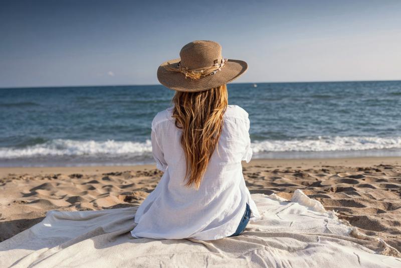 Une femme assise sur une couverture à la plage, regardant la mer, portant un chapeau de paille et une chemise blanche.